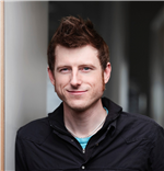 Portrait of Dylan Glosecki smiling at the camera wearing a dark button up shirt in front of a grey background