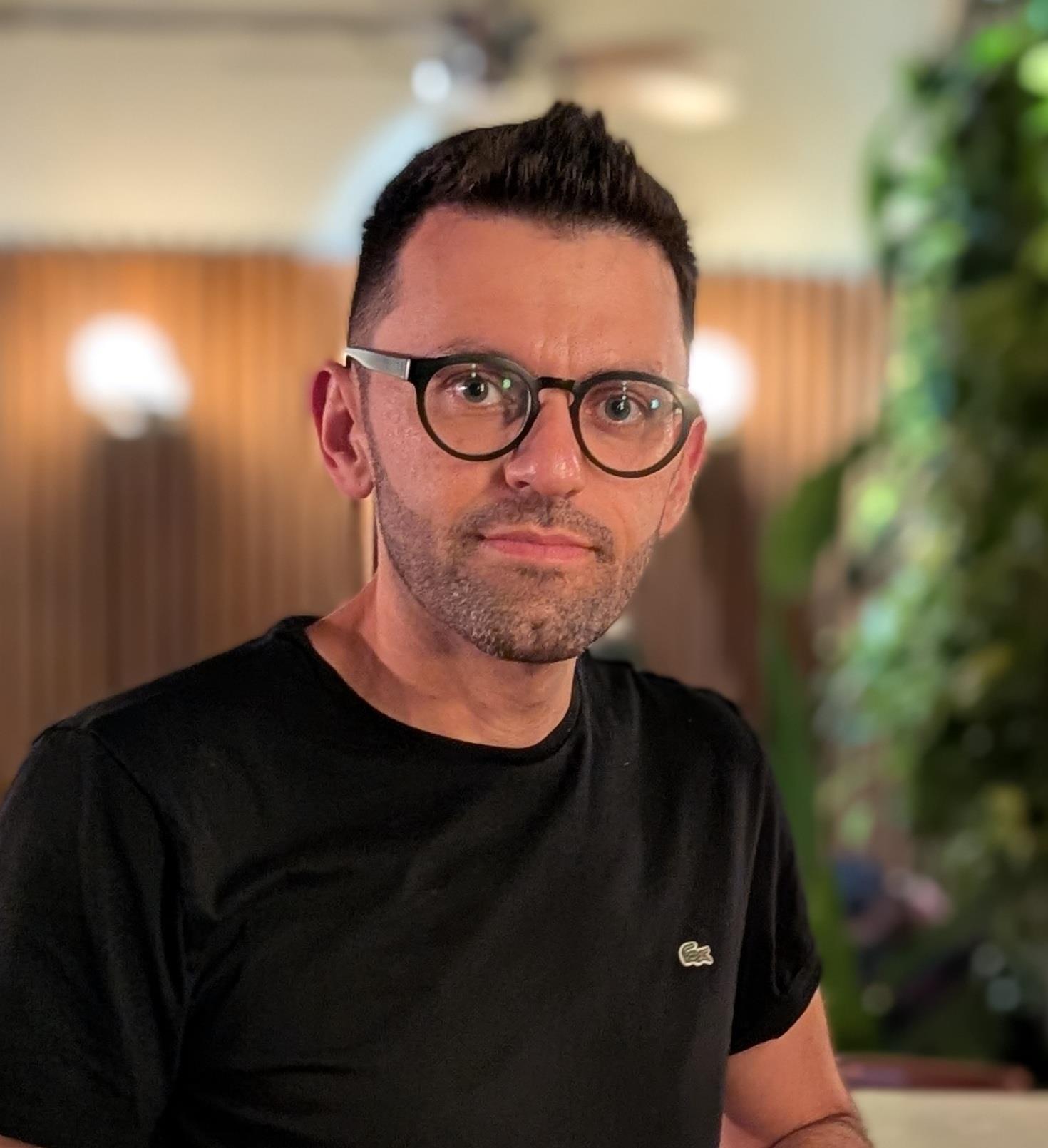 Portrait of Amir smiling slightly at the camera in a dark shirt in front of a wall with wood paneling and greenery