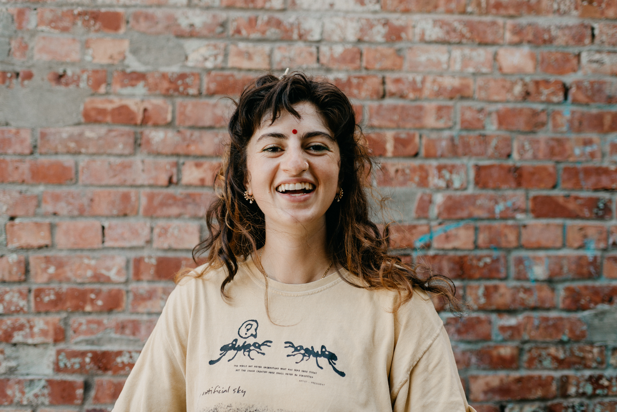 Portrait of Monika smiling wide at the camera in a yellow t-shirt in front of a brick background.