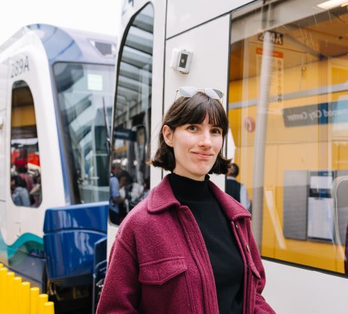 Portrait of Rebecca smiling slightly at the camera in a magenta coat with a light rail train in the background