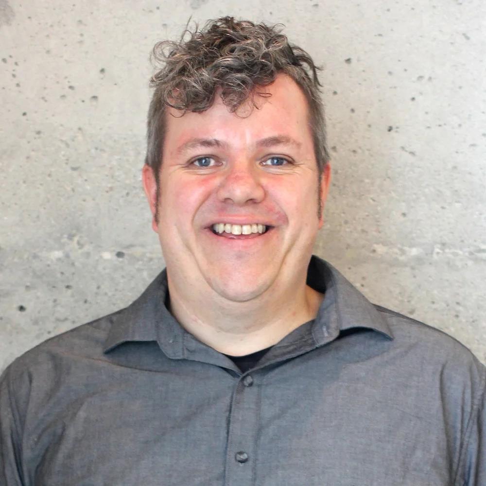 Portrait of Patrick smiling broadly at the camera in a grey shirt in front of a sone background